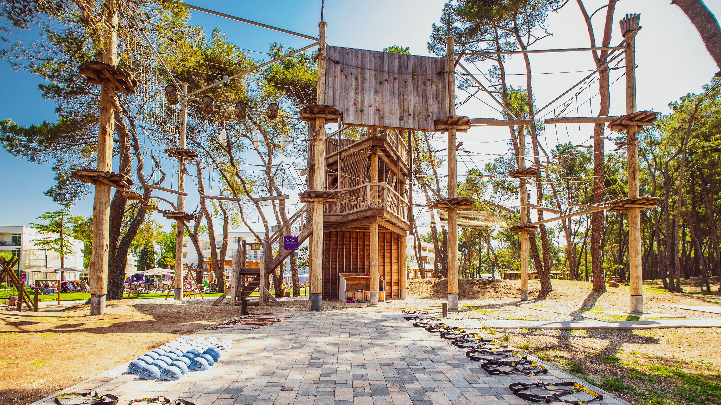 A wooden structure with rope bridges and climbing holds, surrounded by trees and a paved path.