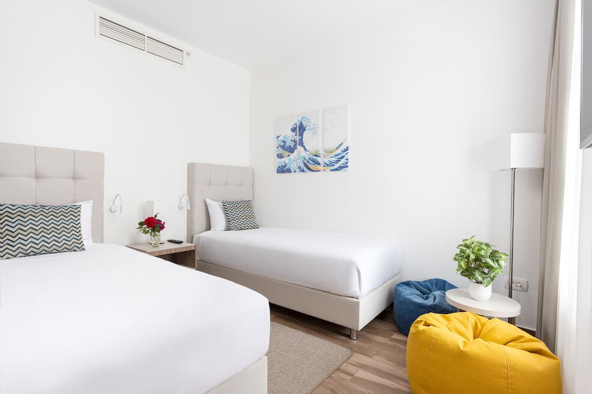 Een hotelkamer met twee bedden, een nachtkastje met een bloevas, een kunstwerk aan de muur, een vloerlamp en een klein, rond tafeltje met een plant.