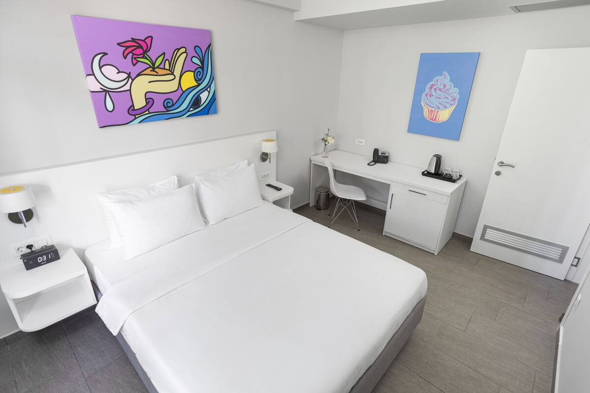 Een moderne hotelkamer met een groot bed, een bureau en een stoel. Er hangt een schilderij aan de muur en een deur met een ventilatie rooster.