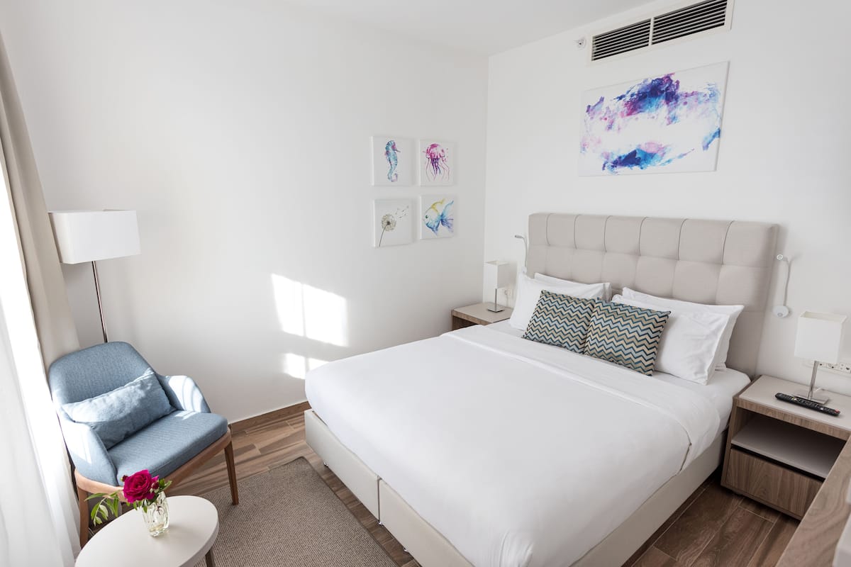 Een moderne hotelkamer met een groot bed, een stoel, een klein tafeltje met een vaas, en kunstwerken aan de muur.