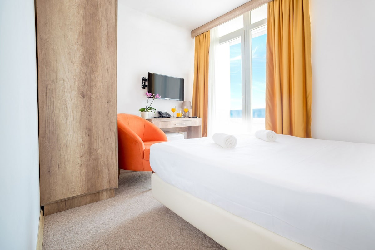 Een hotelkamer met een bed, een bureau met een computer, een oranje fauteuil, een televisie en een raam met gordijnen.
