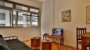 Una habitación con una cama, un televisor sobre un mueble de madera, una mesa de comedor con sillas y una ventana con persianas.