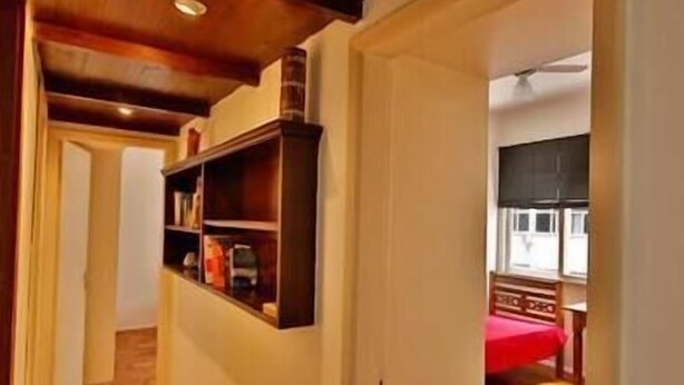 Un estrecho pasillo con piso de madera, una estantería empotrada y una puerta que da a una habitación con cama y ventana.