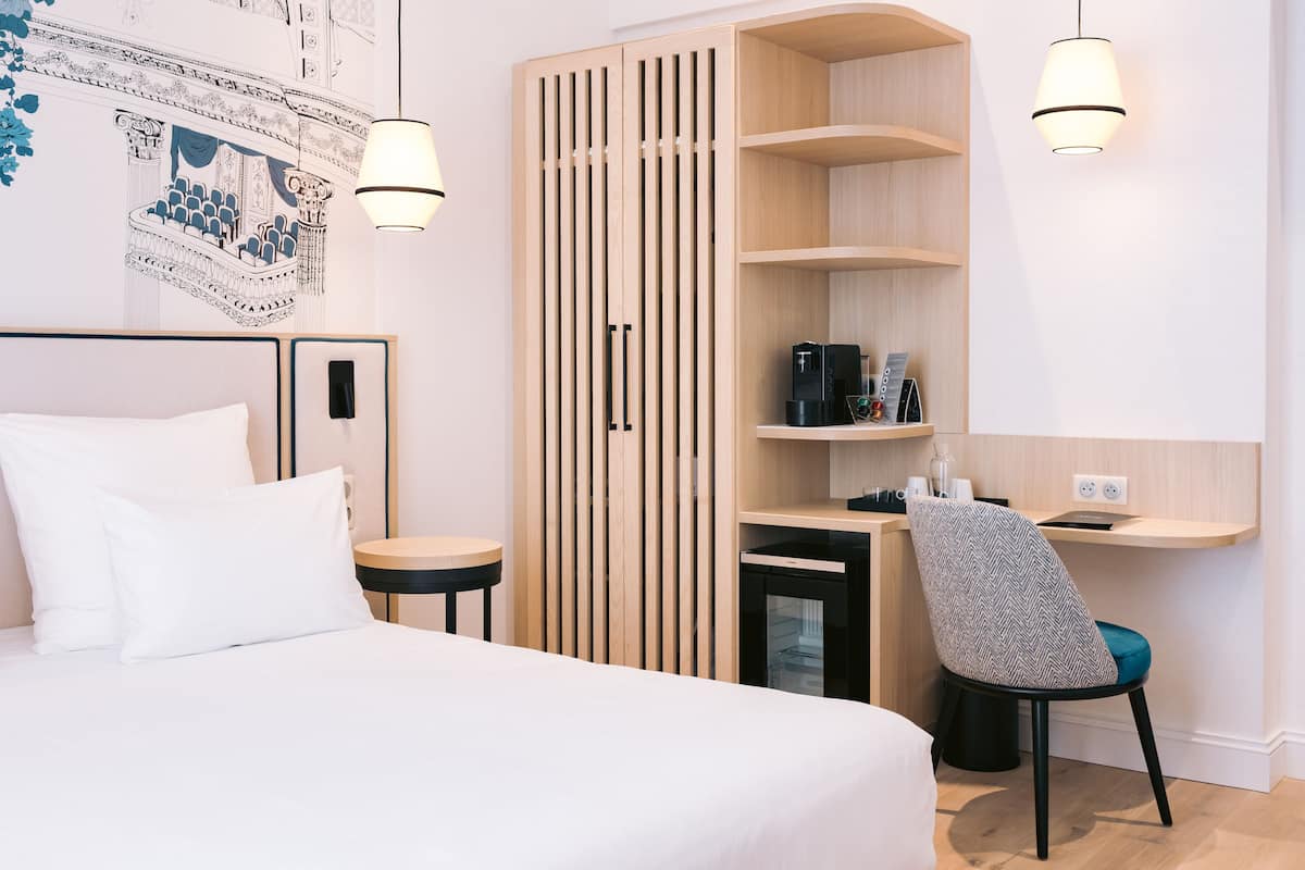 A modern hotel room with a bed, a desk, a chair, a wardrobe, and a coffee machine.