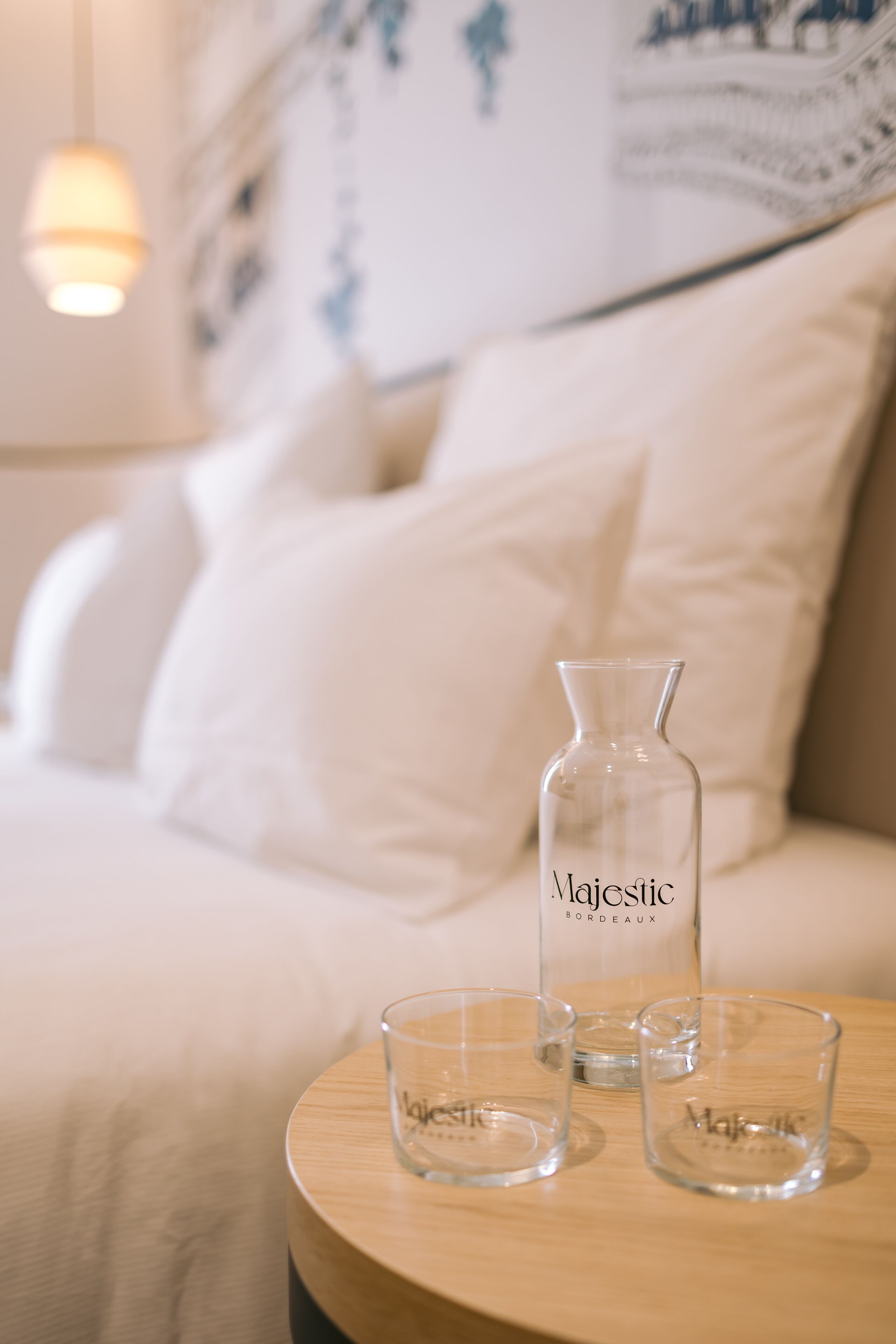 A bed with white bedding and pillows, a bedside table with a clear bottle labeled "Majestic Bordeaux" and two glasses.