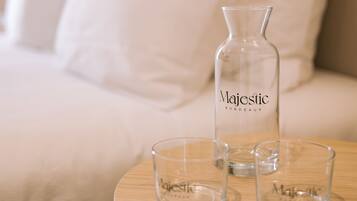 A bed with white bedding and pillows, a bedside table with a clear bottle labeled "Majestic Bordeaux" and two glasses.