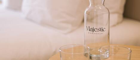 A bed with white bedding and pillows, a bedside table with a clear bottle labeled "Majestic Bordeaux" and two glasses.