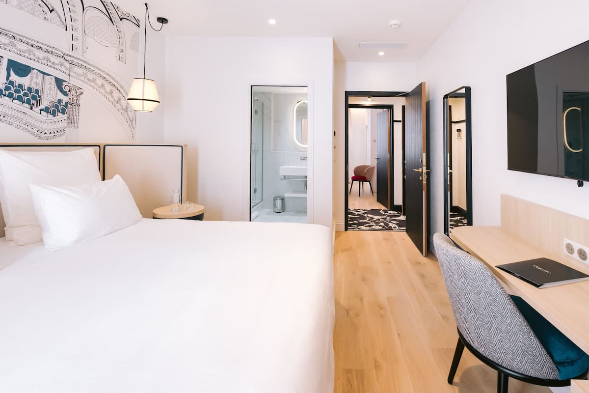 A modern hotel room with a large bed, a desk with a chair, a mirror, and a wall-mounted TV.