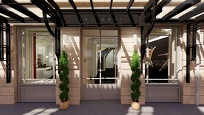 La entrada del Hotel Majestic, con un diseño moderno, presenta un frente de vidrio, una escalera y dos árboles de hoja perenne en macetas.