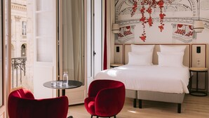 Habitación de hotel con una cama grande, dos sillas rojas, una mesita y vistas a la ciudad a través de una puerta abierta.