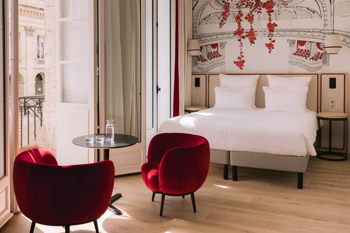 A hotel room with a large bed, two red chairs, a small table, and a view of the city through an open door.