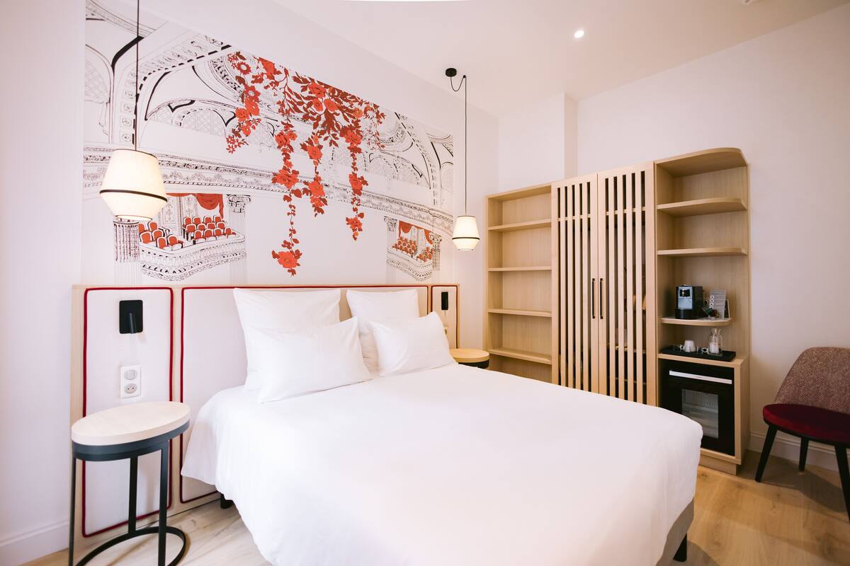 A hotel room with a large bed, a side table, a chair, a wooden bookshelf, and a wall with a decorative map.