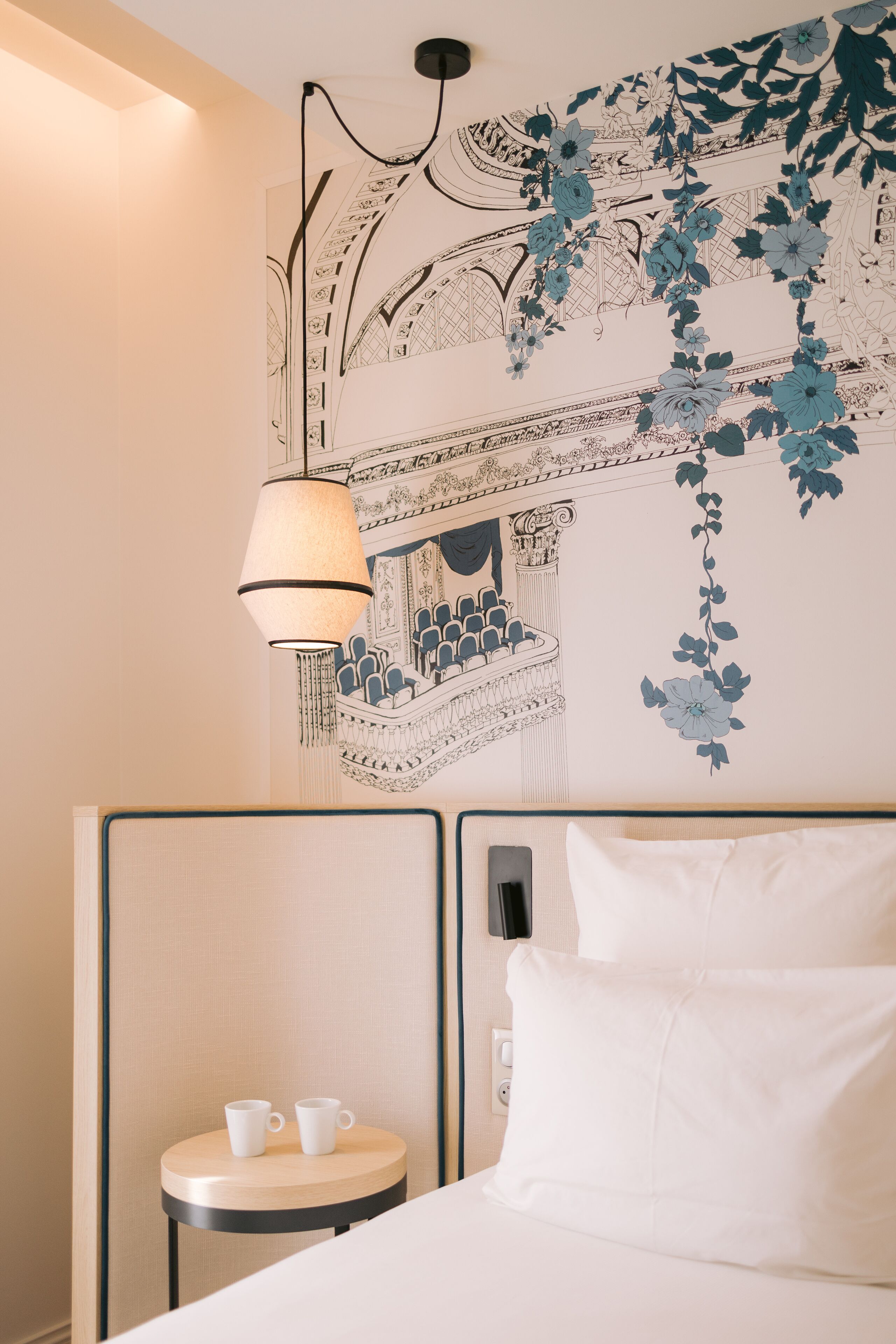 Une chambre d’hôtel avec un lit, une petite table ronde sur laquelle sont posées deux tasses blanches, une lampe fixée au mur et une fresque décorative murale.