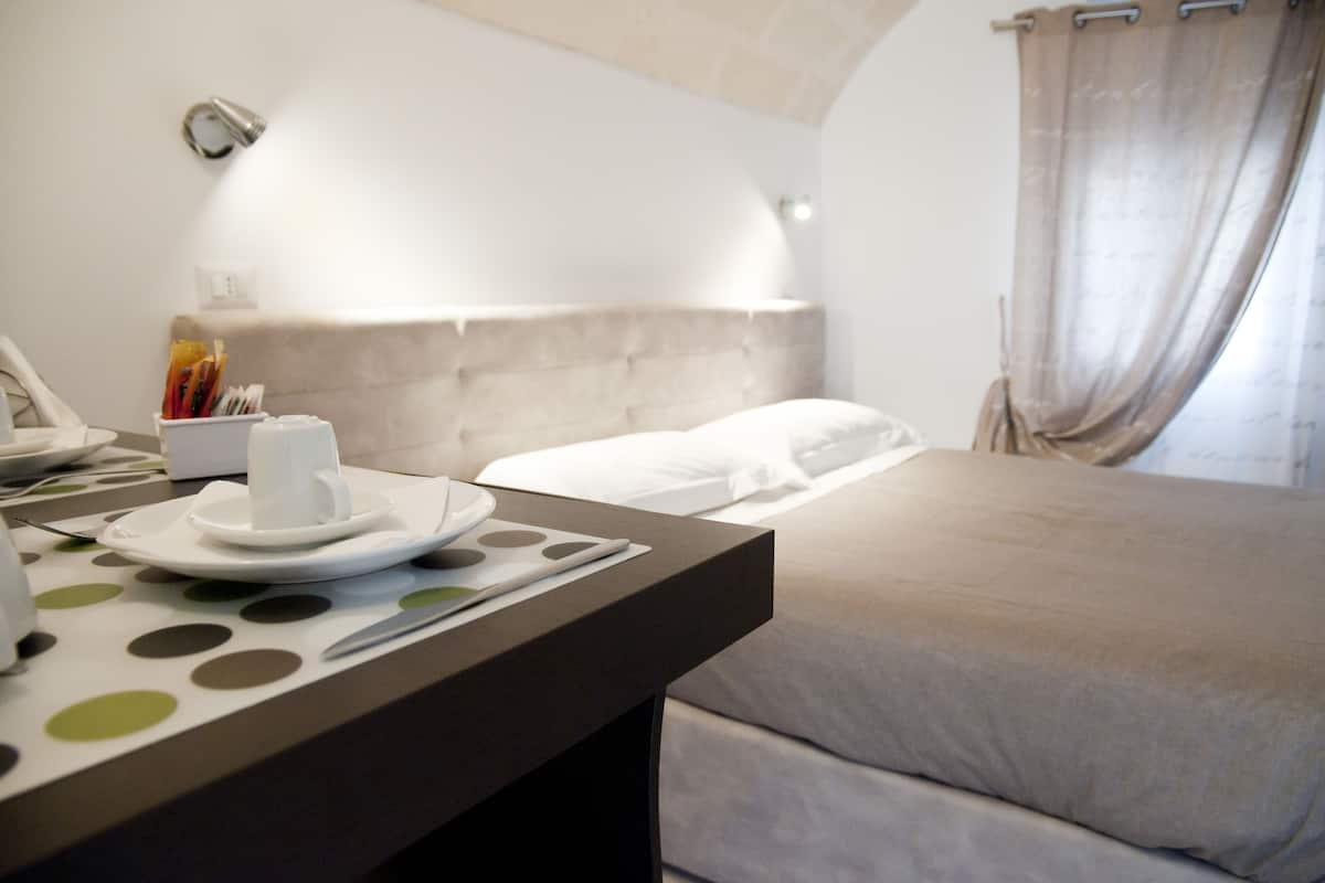 A hotel room with a bed, a bedside table with a cup and saucer, a phone, and a small table with a polka dot tablecloth.