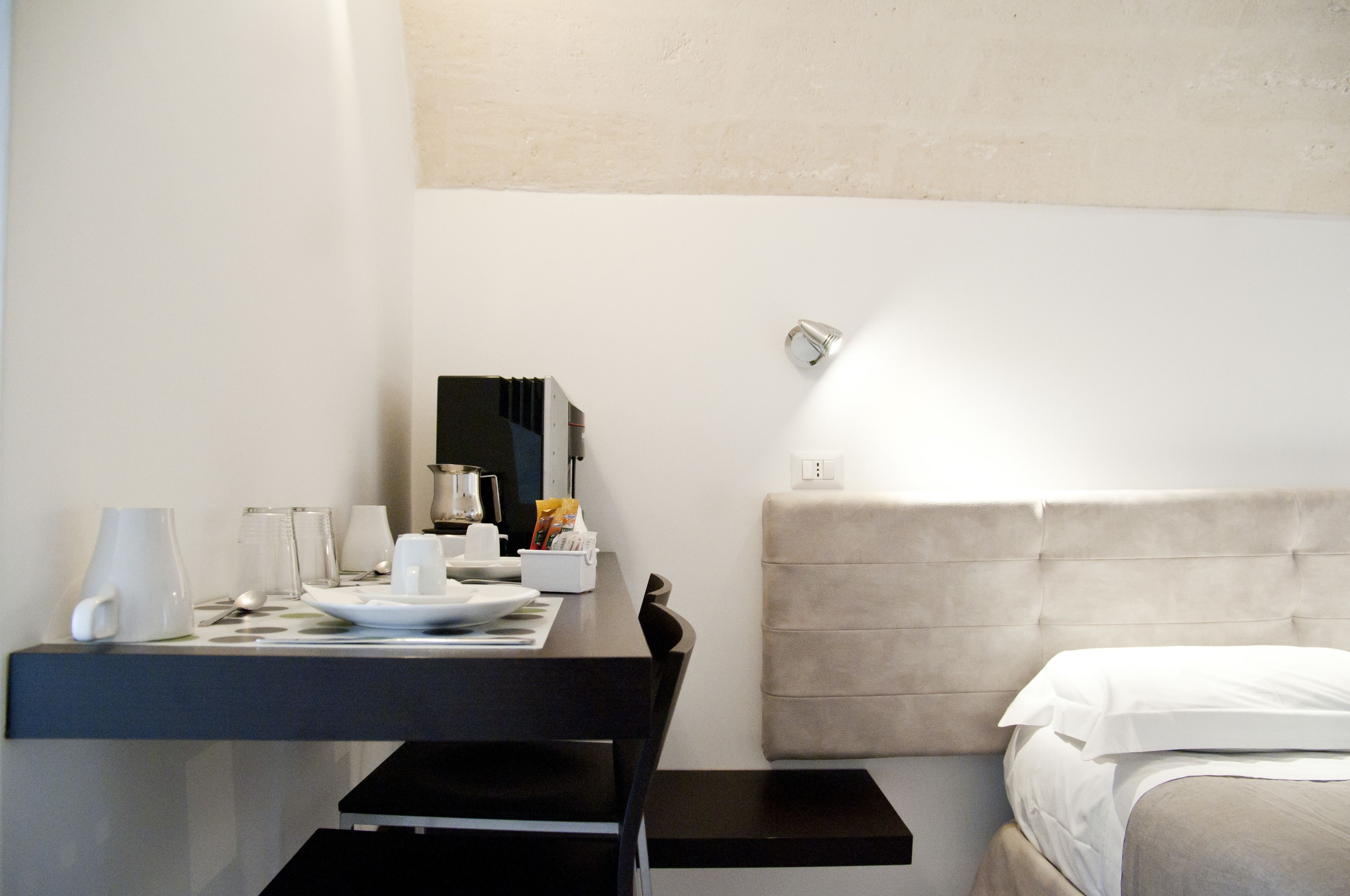 A hotel room with a bed, a bedside table with a lamp, a small table with a coffee maker, and a mirror on the wall.