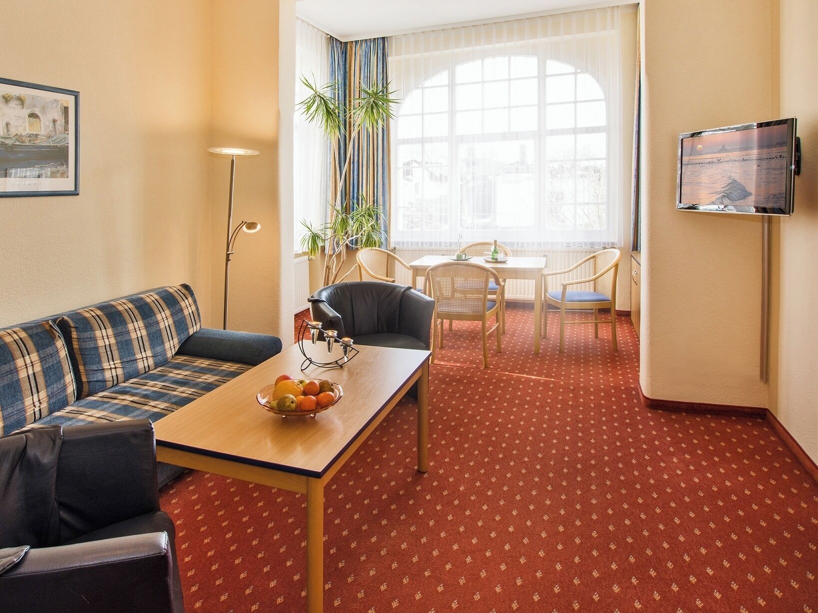 Ein Hotelzimmer mit rotem Teppich, einer Couch, einem Esstisch mit Stühlen, einem Fernseher und einem großen Fenster mit Vorhängen.
