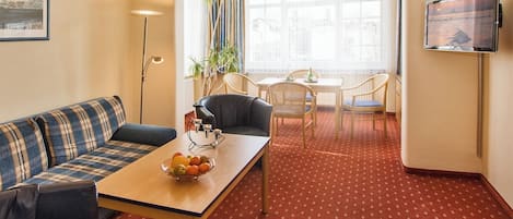 Ein Hotelzimmer mit rotem Teppich, einer Couch, einem Esstisch mit Stühlen, einem Fernseher und einem großen Fenster mit Vorhängen.