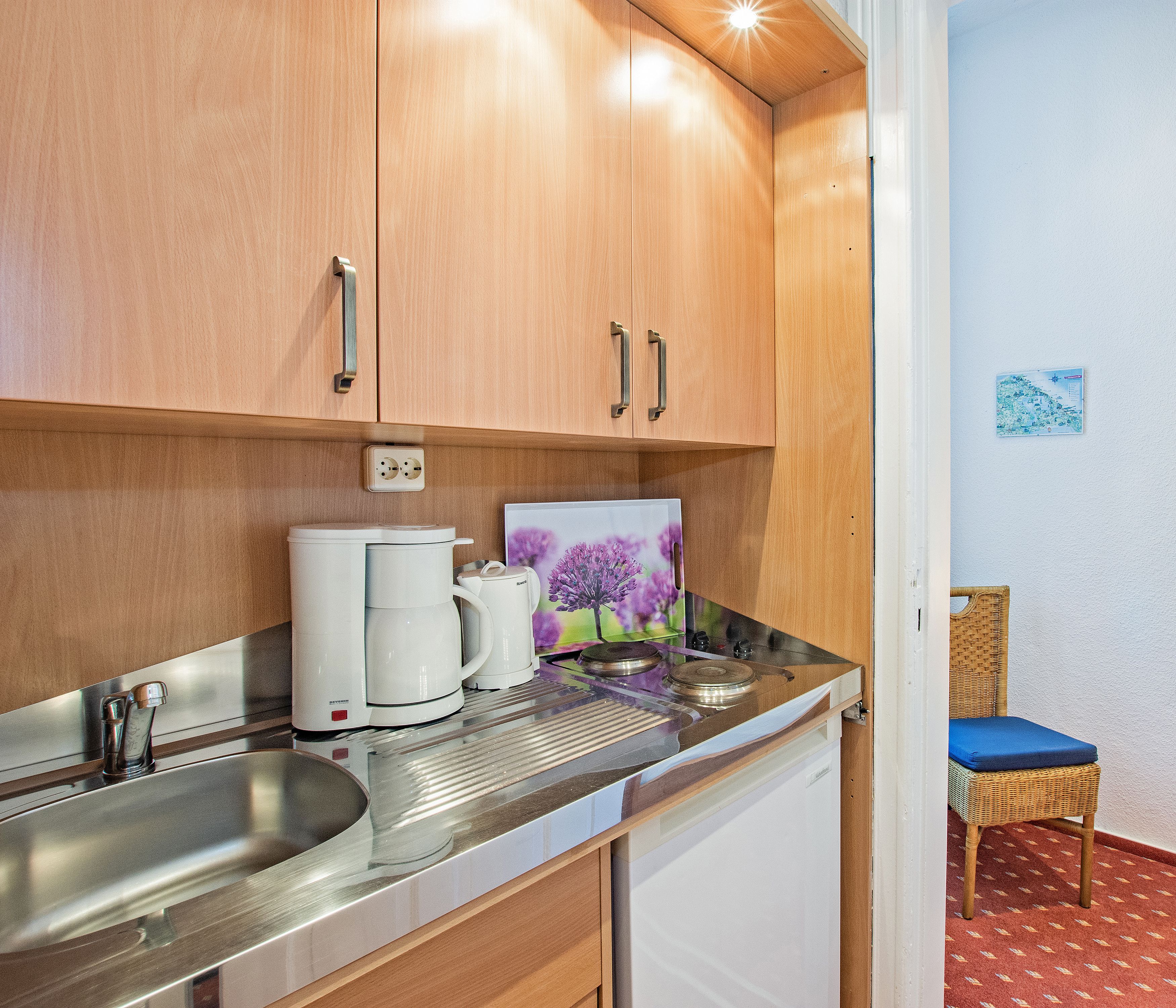 Een kitchenette met houten kastjes, een roestvrijstalen spoelbak en een kleine koelkast. Op het aanrecht staat een afbeelding van paarse bloemen.
