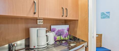 Een kitchenette met houten kastjes, een roestvrijstalen spoelbak en een kleine koelkast. Op het aanrecht staat een afbeelding van paarse bloemen.
