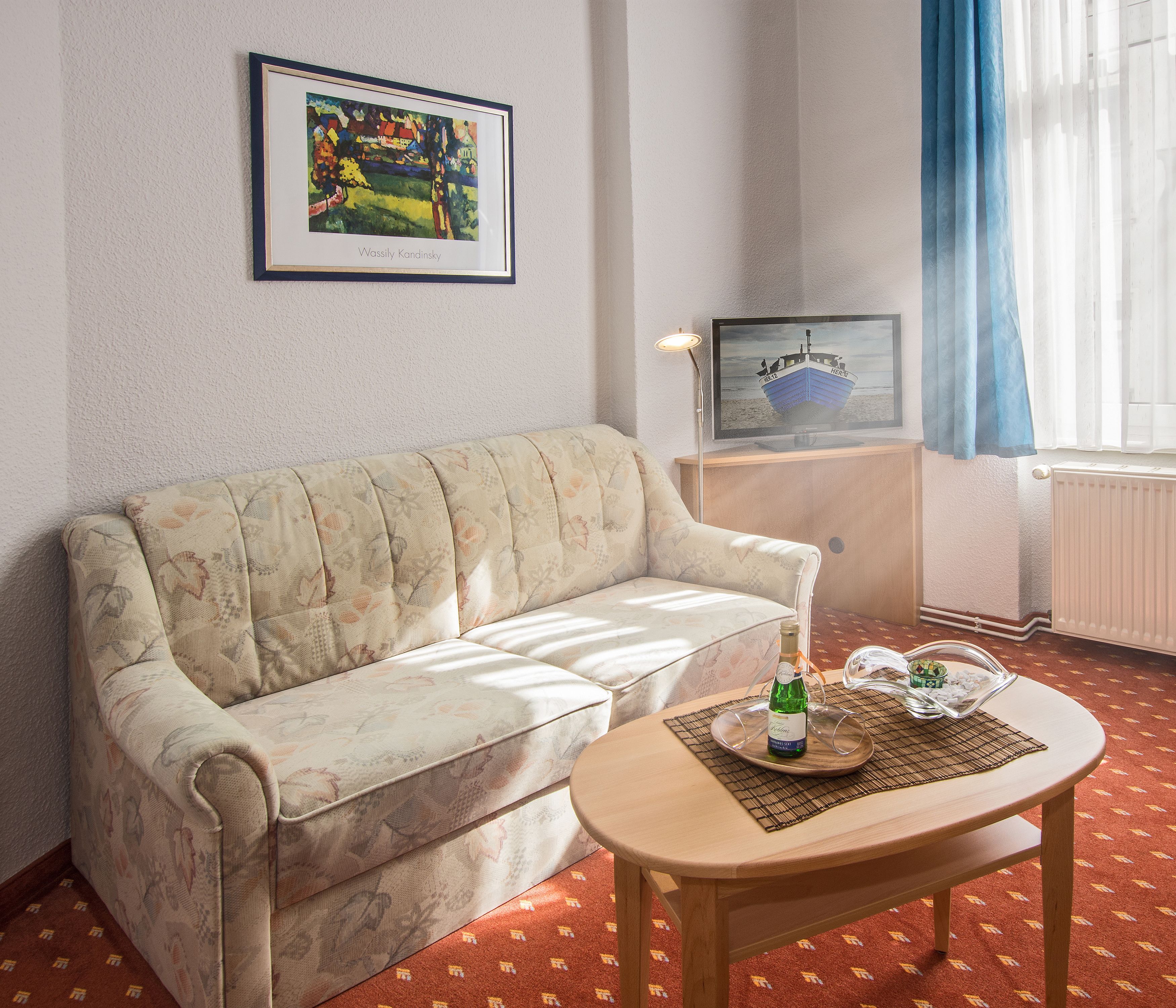 A living room with a sofa, a coffee table with a bottle and glasses, a framed painting, and a boat painting on the wall.