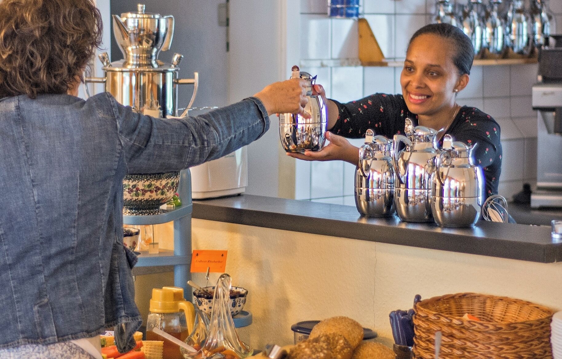 A person holding a silver teapot at a counter with various kitchen items and a sign that reads "Coffee Roasters".