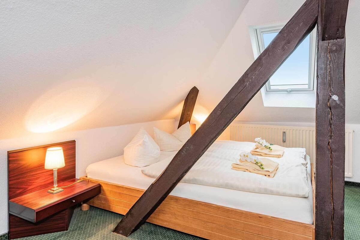A bedroom with a wooden bed, white bedding, a nightstand with a lamp, and a window with a view of the sky.