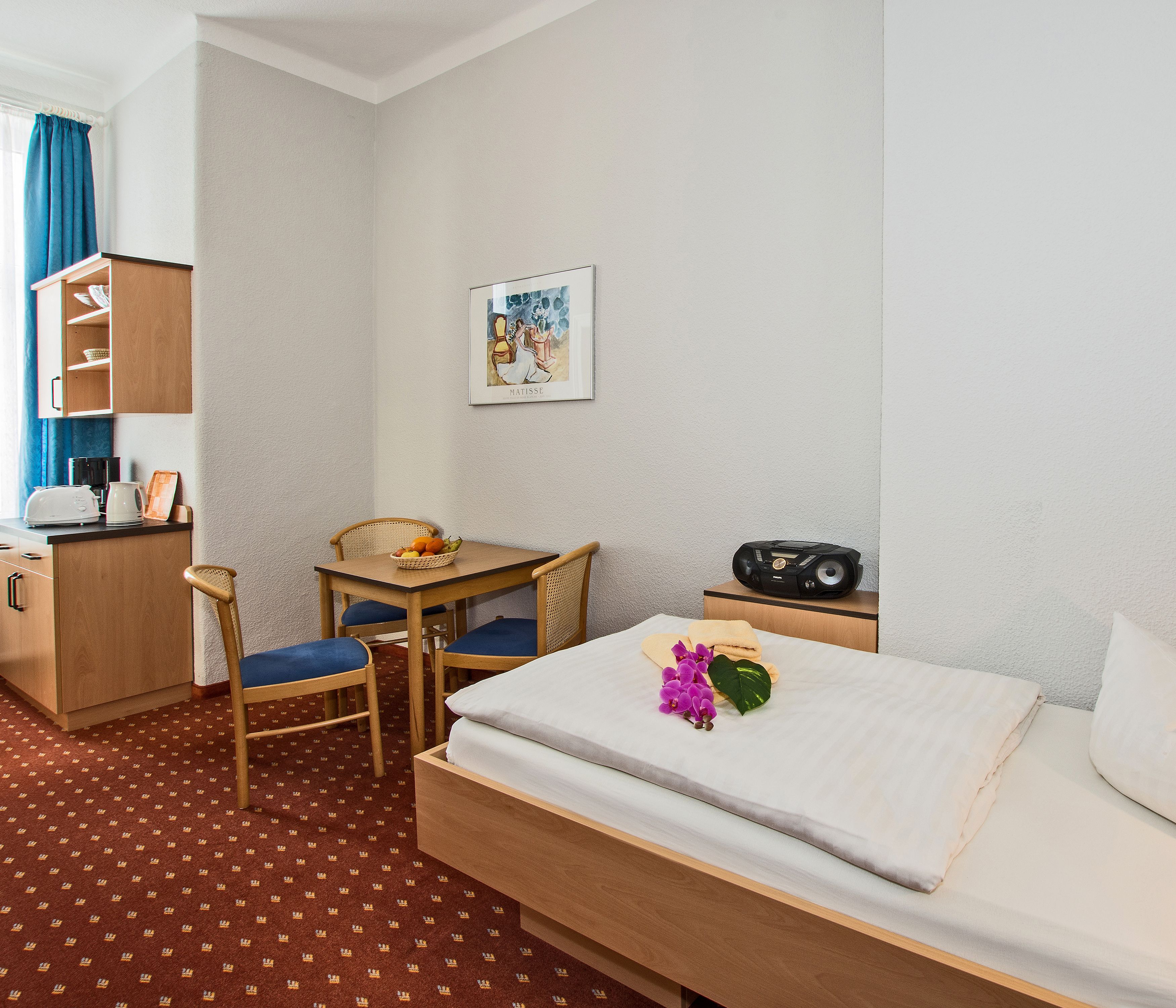 Habitación de hotel con cama, escritorio y silla, una mesita con una cesta de fruta, radio y un cuadro en la pared.
