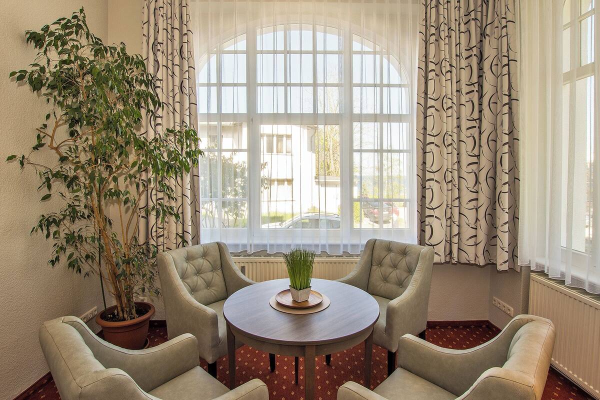 A room with a round table, four armchairs, a potted plant, and a large window with curtains.