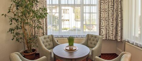 A room with a round table, four armchairs, a potted plant, and a large window with curtains.