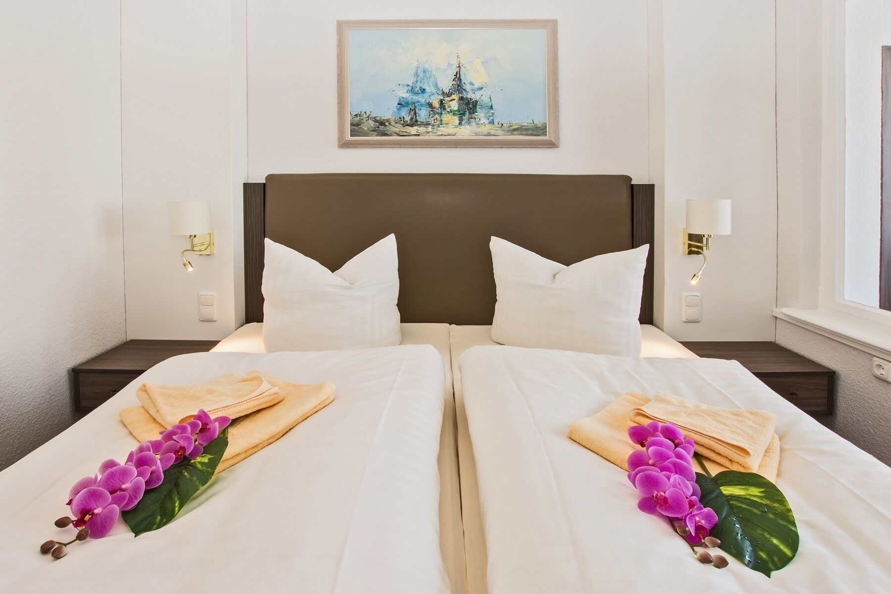 Ett hotellrum med två sängar, vita sängkläder och handdukar. På sängarna finns dekorativa, lila orkidéer och ett grönt blad. En tavla hänger på väggen ovanför sängarna.
