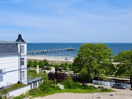 Vista costera con un muelle que se adentra en el mar, una playa y un edificio con balcón con vistas al agua.