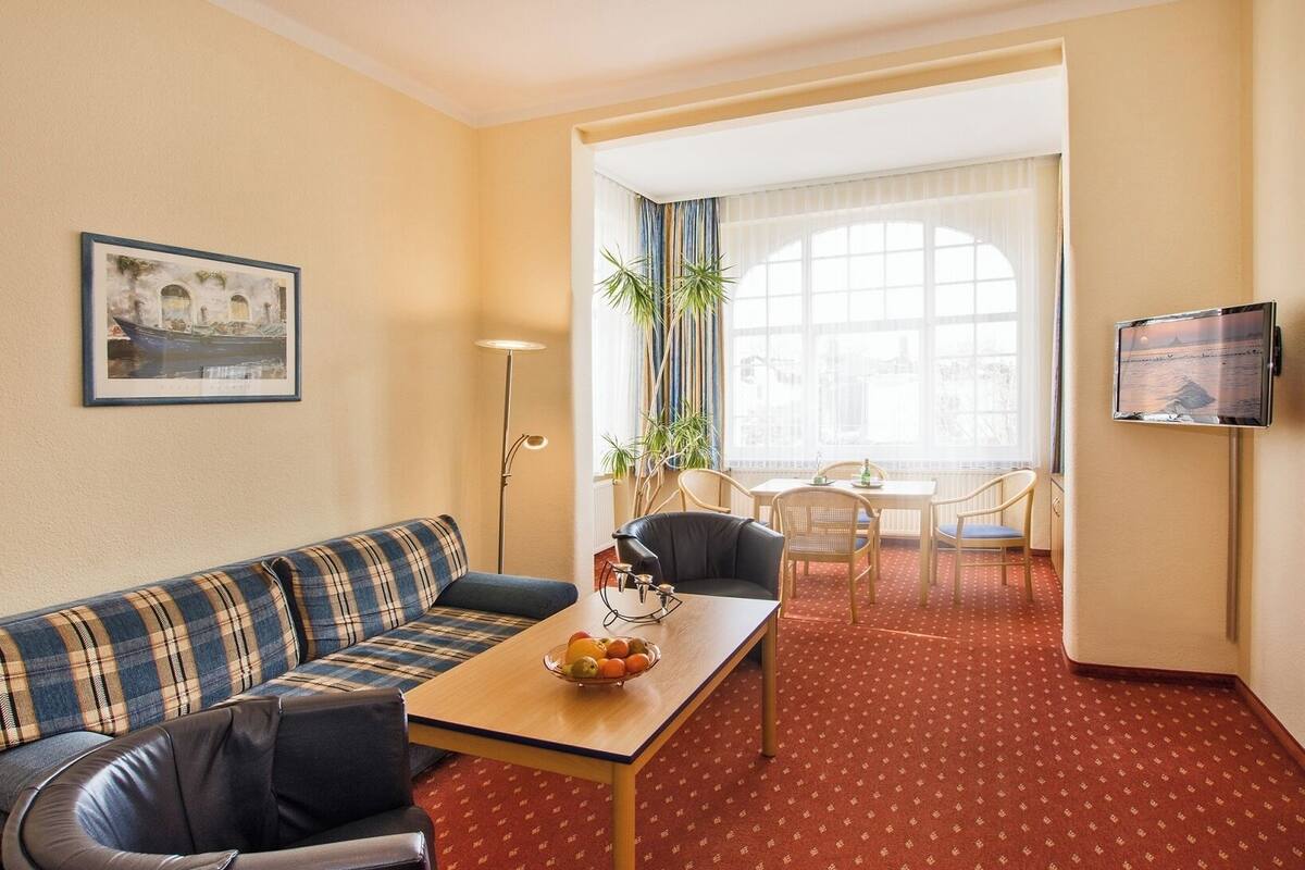 A hotel room with a sofa, a dining table with fruit, a TV, and a view of the city through large windows.