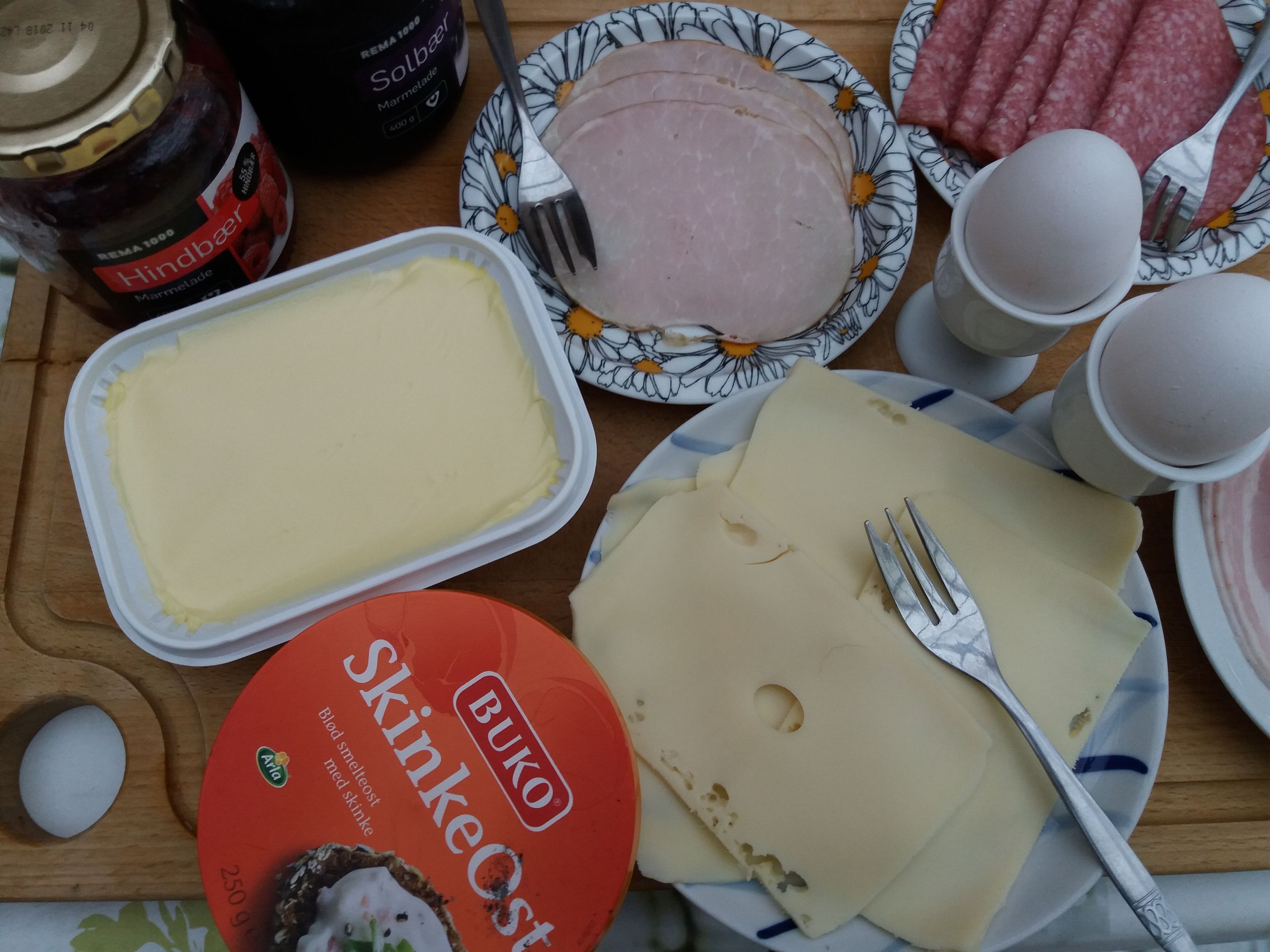 Une table avec différents aliments, notamment une assiette de jambon, un pot de crème, une assiette de fromage et une boîte de Skinkeost.