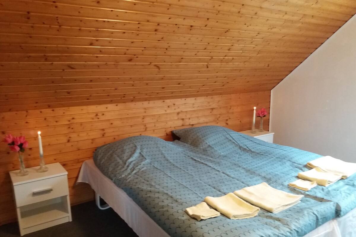 A bedroom with a sloped ceiling, wooden walls, a bed with a blue patterned bedspread, two bedside tables with candles, and a small white nightstand.