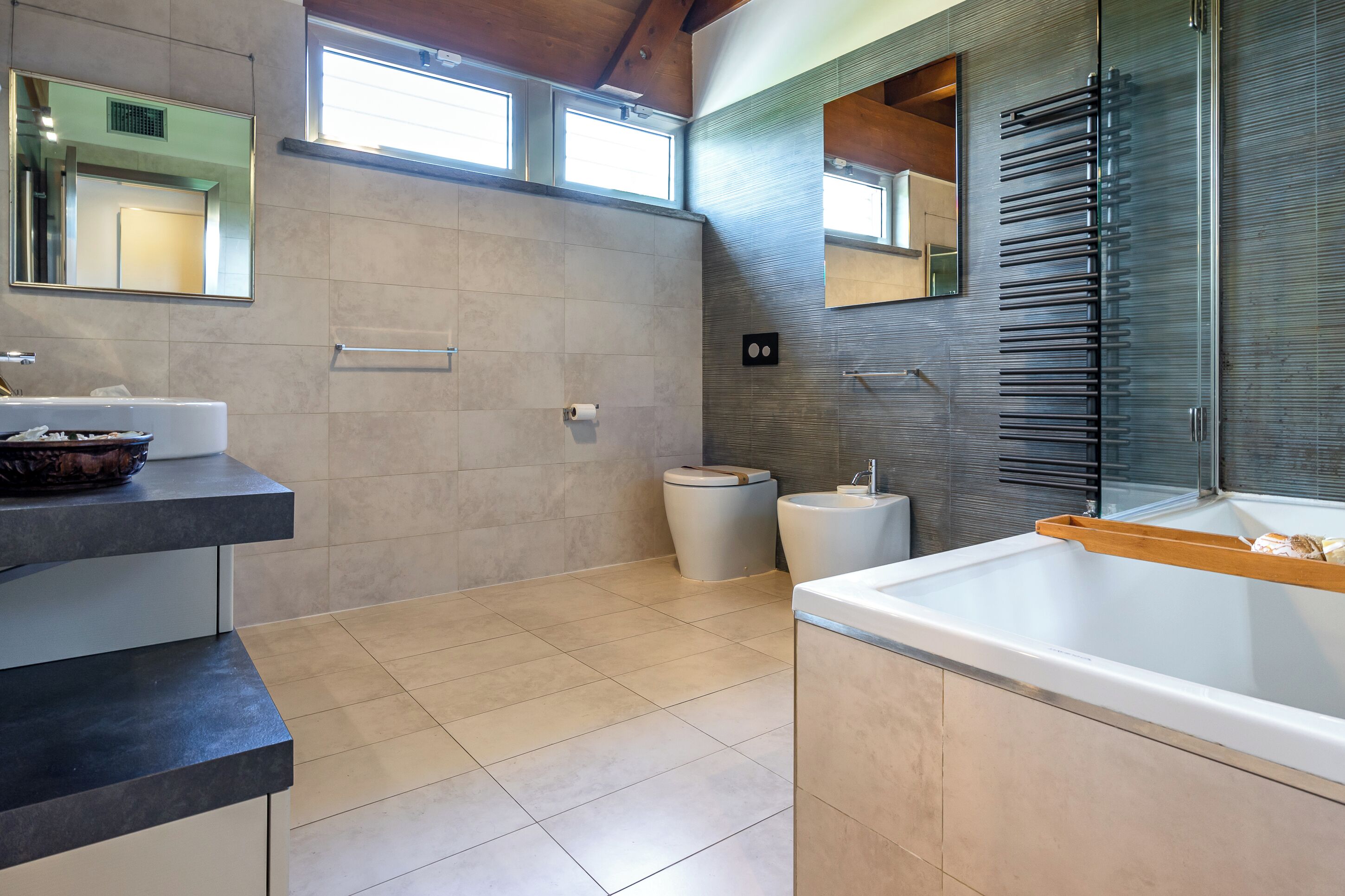 Ein modernes Badezimmer mit einer Badewanne, einem Waschbecken und einer Toilette. Fenster sorgen für natürliches Licht, und über dem Waschbecken befindet sich ein Spiegel.