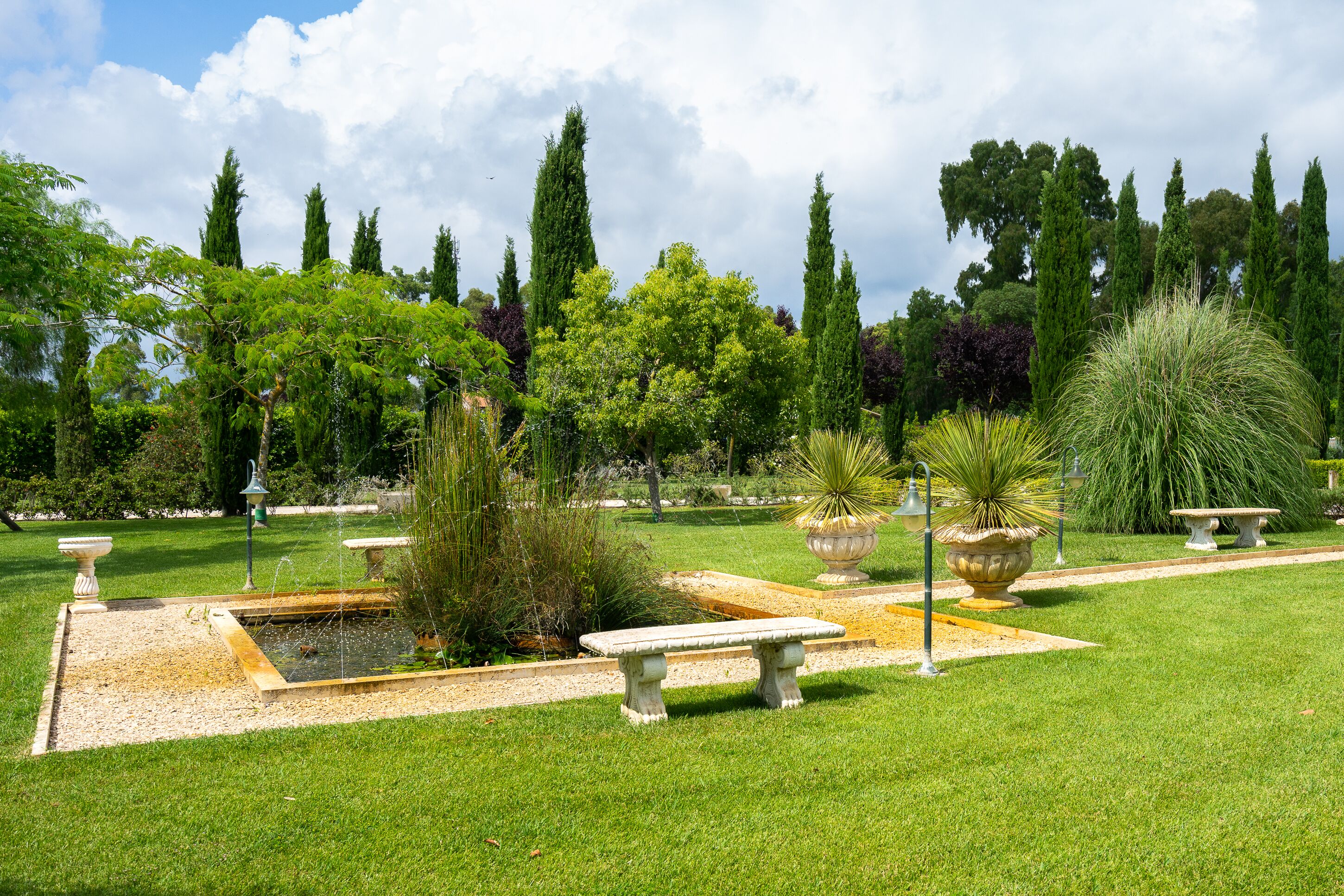 Un jardín bien cuidado con una fuente central, rodeado de setos ordenados y árboles altos.