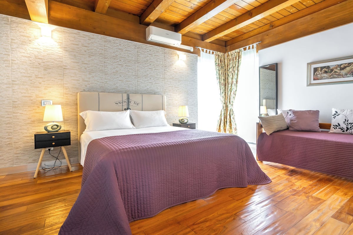 A bedroom with a large bed, a smaller bed, wooden flooring, a stone wall, and a ceiling fan.