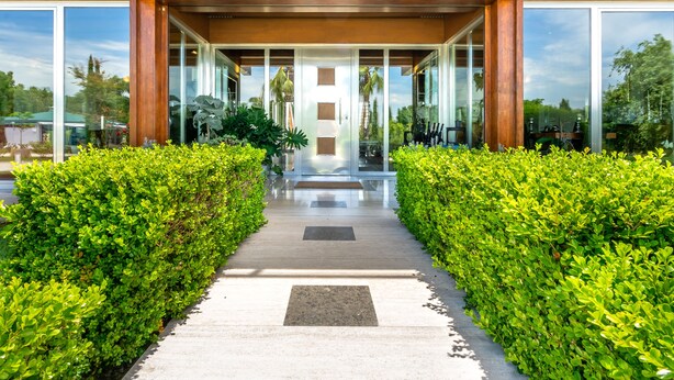A modern building entrance with a glass facade, a concrete pathway, and green hedges on either side. - Casale delle Ronde B&B (Latina)