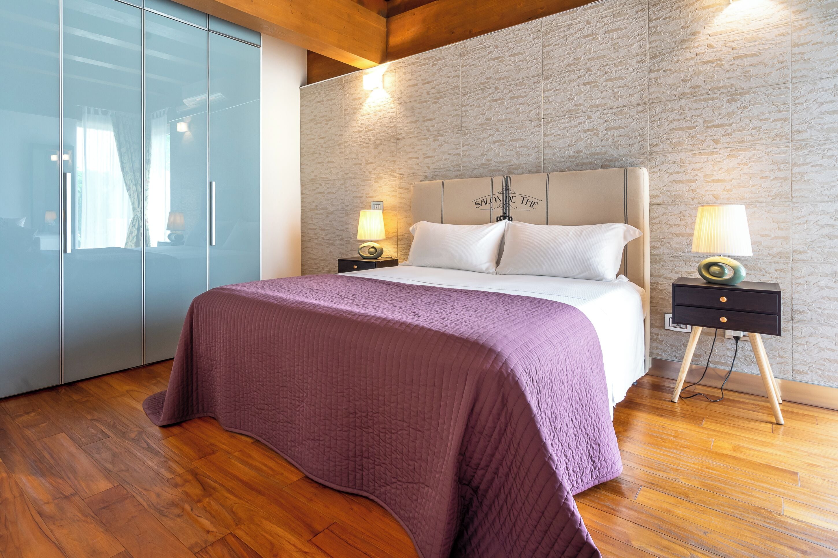 A bedroom with a large bed, two bedside tables with lamps, a wardrobe, and a textured wall.
