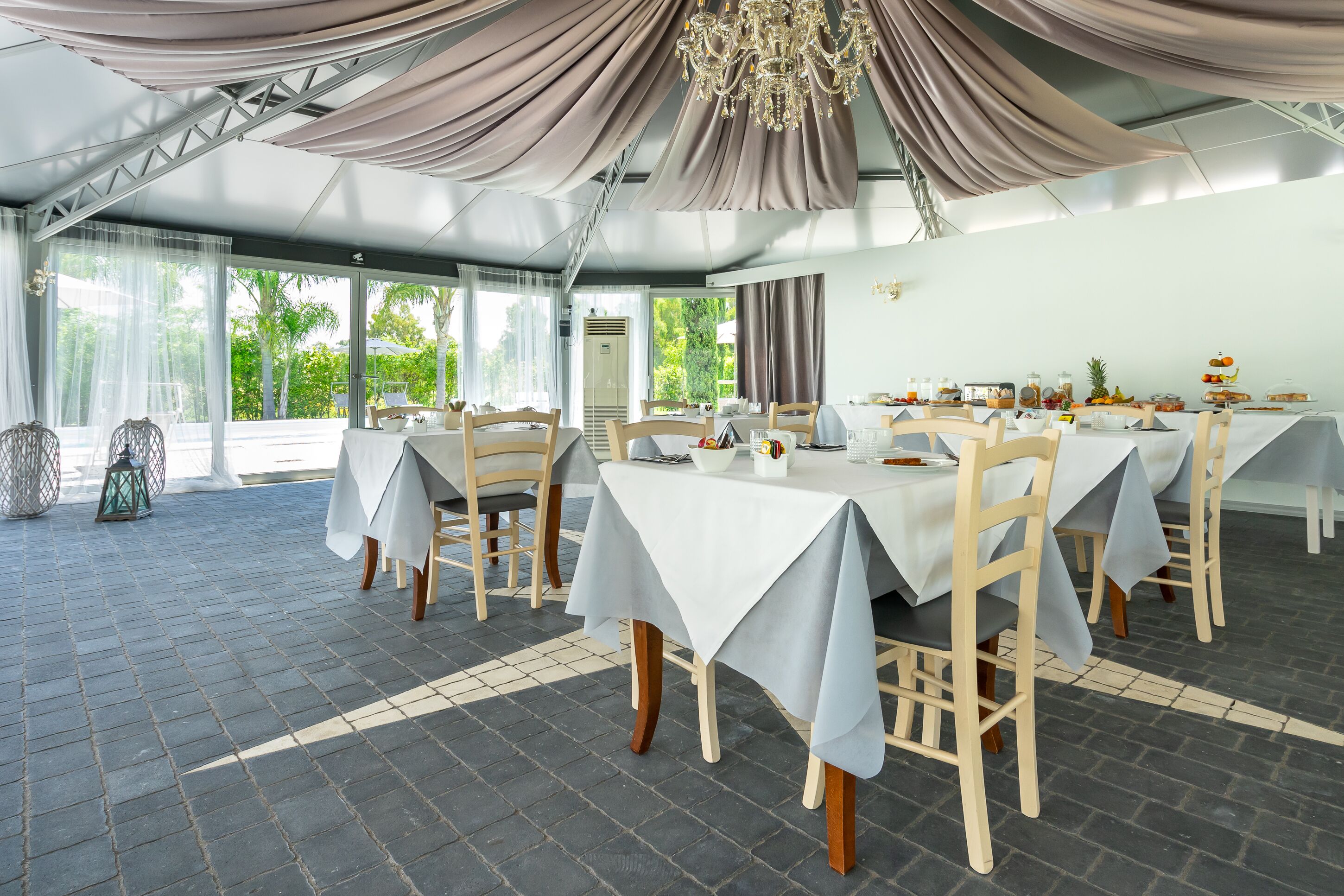 A restaurant with tables set for dining, a chandelier, and a view of greenery outside.