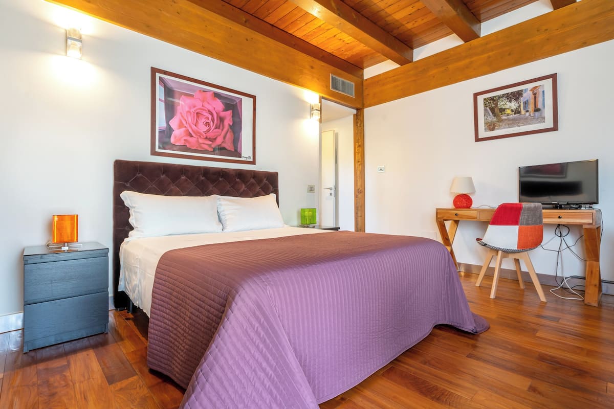 A bedroom with a large bed, a desk with a computer, a TV, and artwork on the walls.