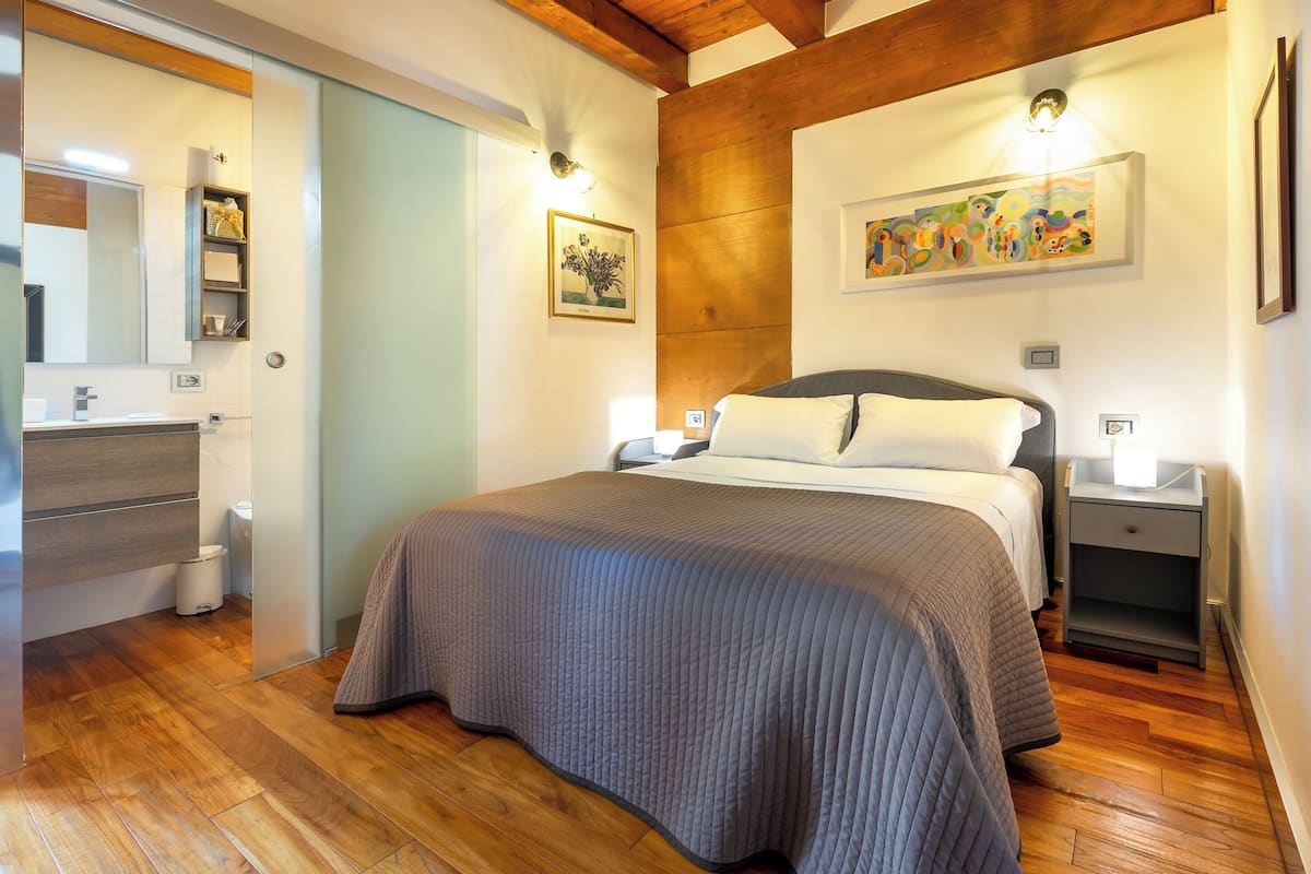 A bedroom with a large bed, wooden flooring, a nightstand, a bedside table, a wall-mounted shelf, a mirror, and a painting.