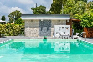 Una piscina con una casa de piscina moderna, sillas de descanso y un muro de piedra.