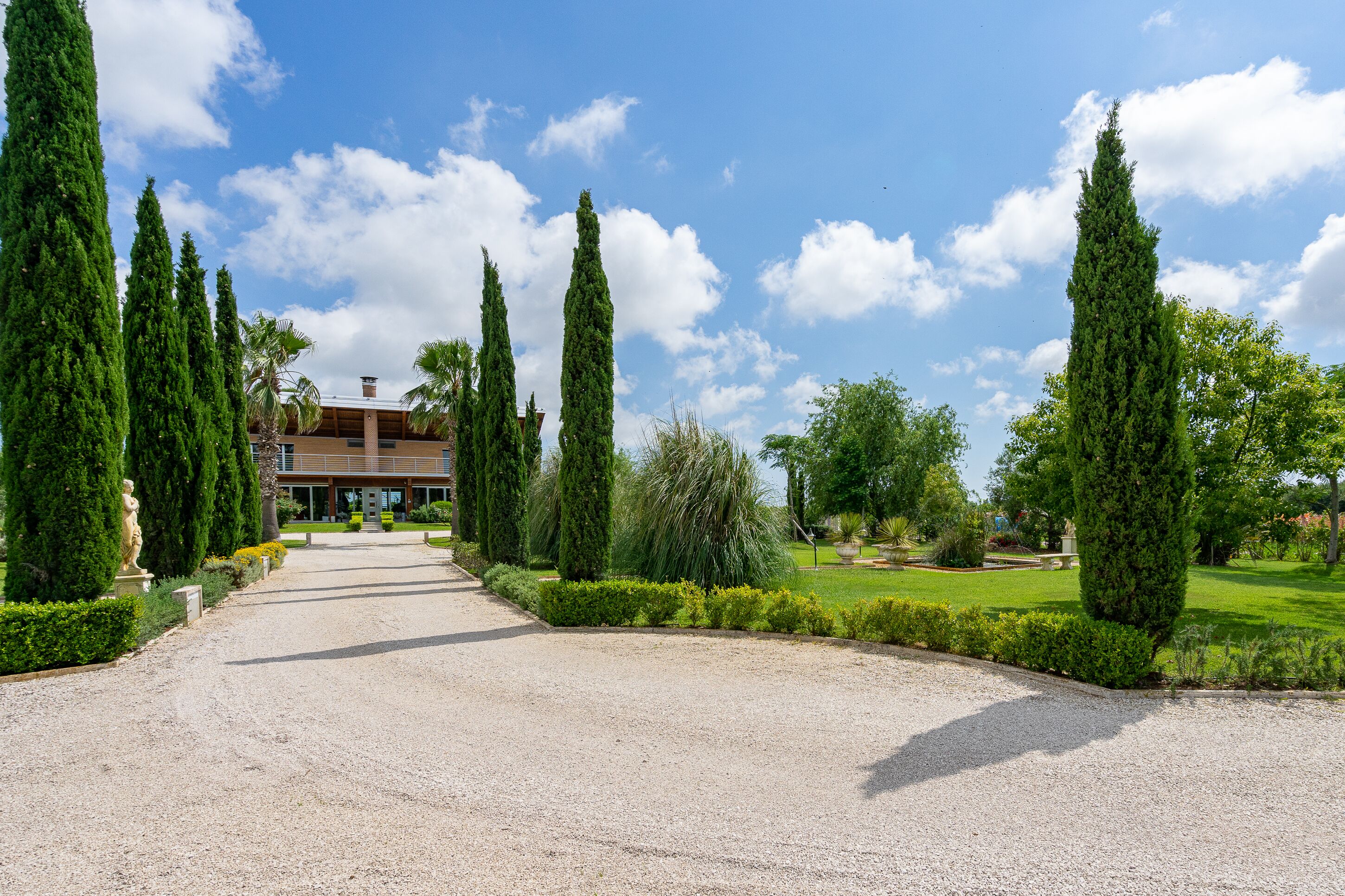 A gravel driveway leading to a building with a modern design, surrounded by neatly trimmed hedges and tall trees.
