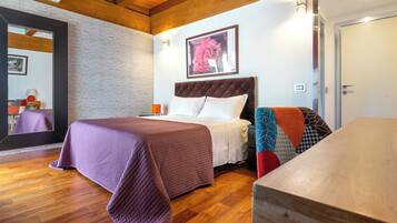 Une chambre avec un plancher en bois, un lit recouvert d’une courtepointe violette, une tête de lit, une table de chevet et un tableau encadré accroché au mur.