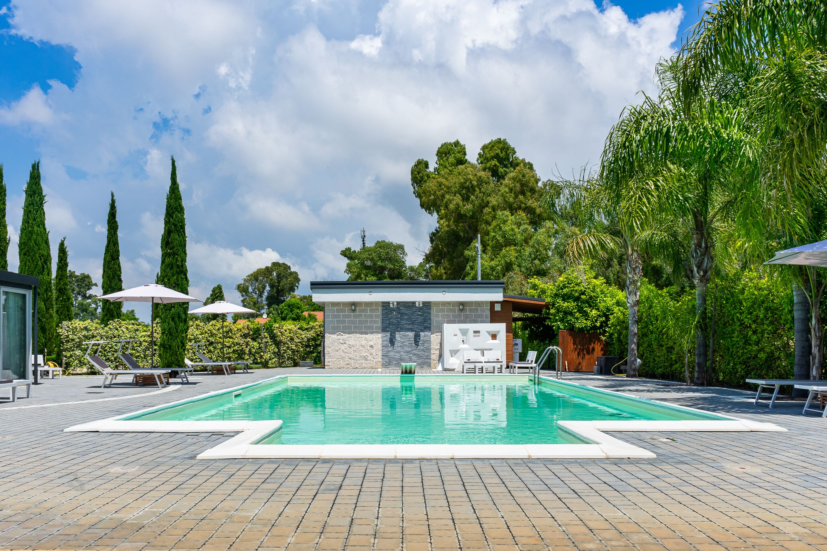 Piscina con sillas de descanso y sombrillas, rodeada de árboles y un muro de piedra.