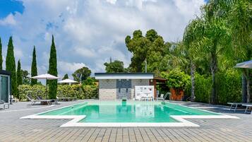 Piscina con sillas de descanso y sombrillas, rodeada de árboles y un muro de piedra.