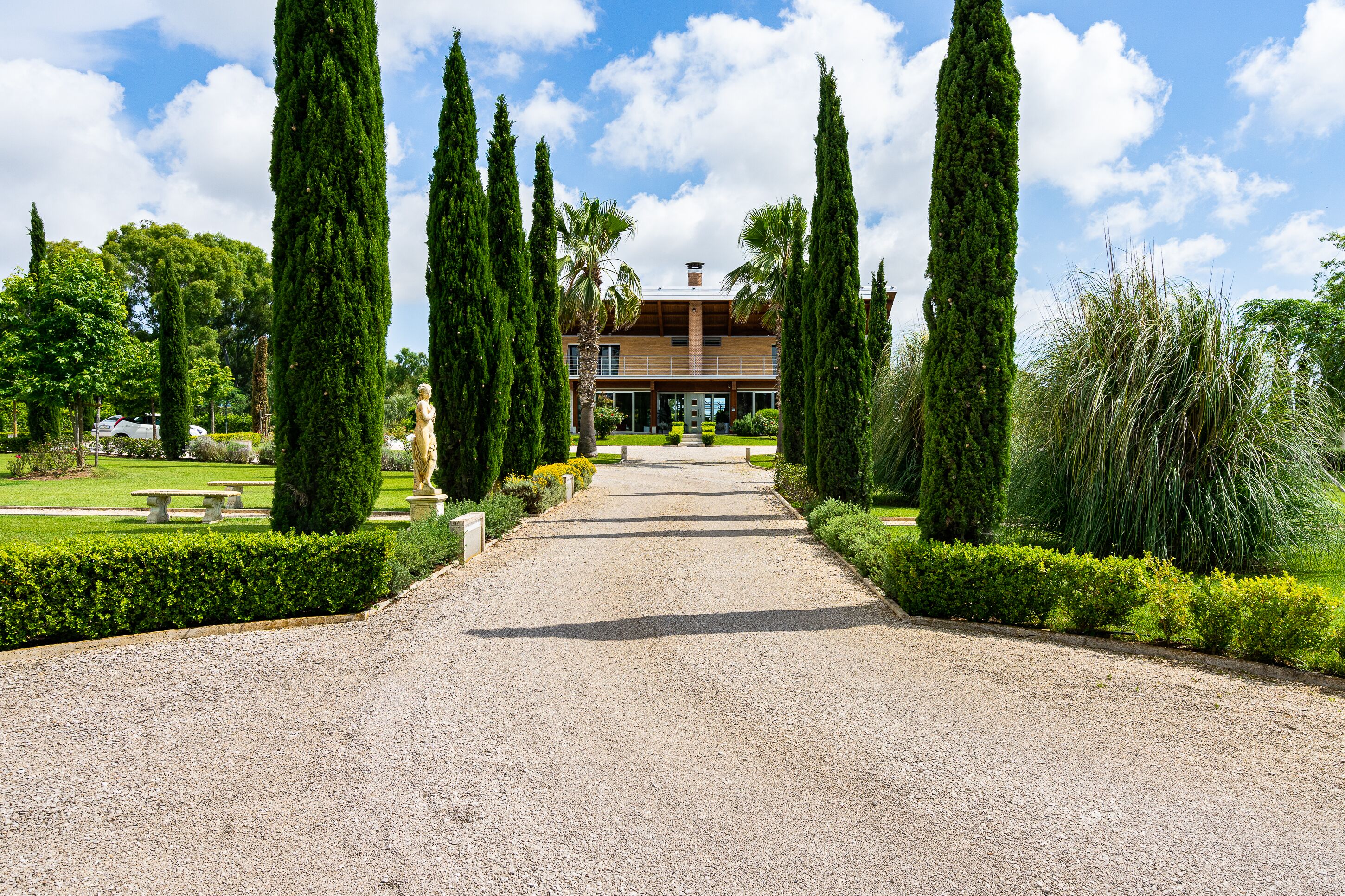 A gravel pathway leading to a building with a symmetrical garden featuring tall cypress trees and manicured hedges.