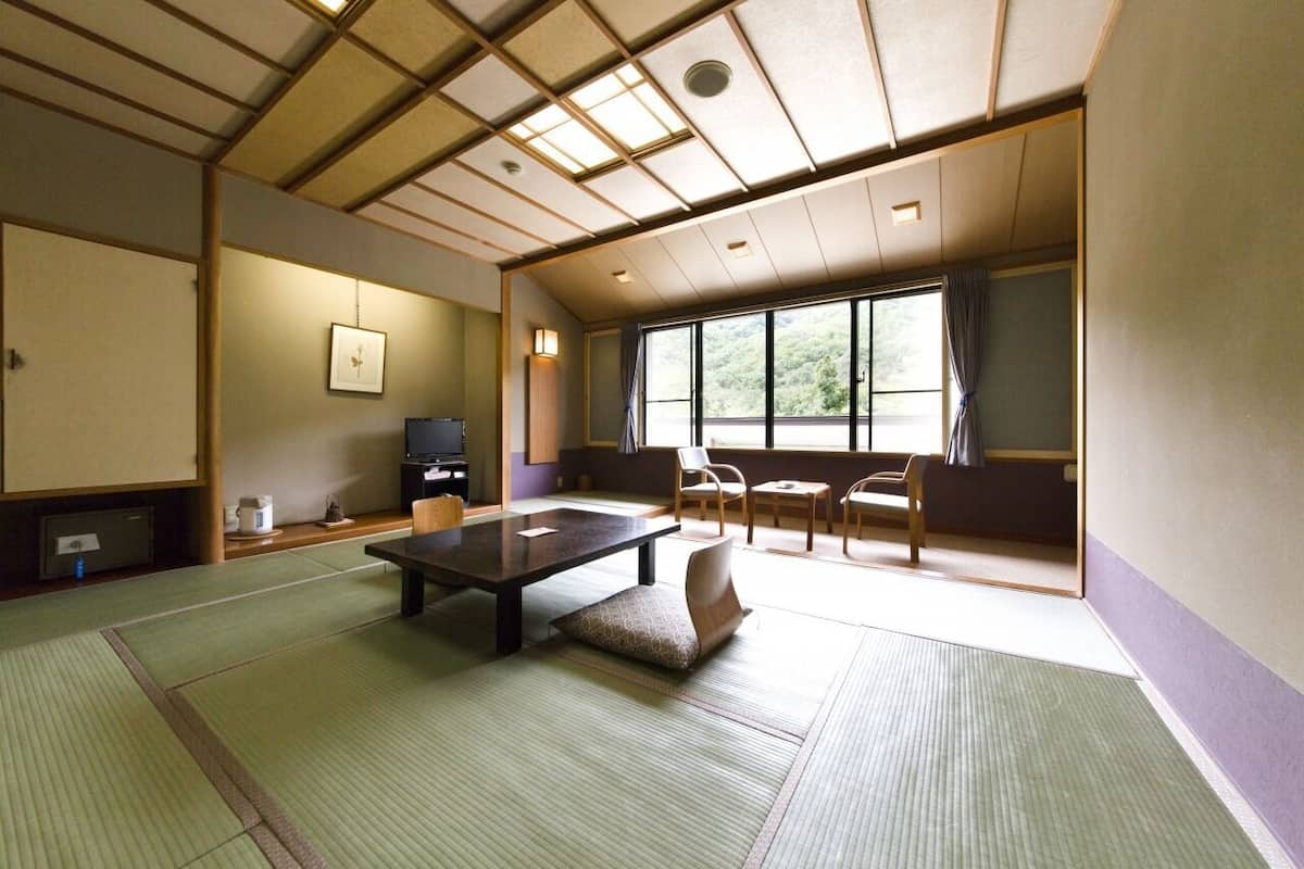 Una habitación japonesa tradicional con suelo de tatami, una mesa baja, sillas, un televisor y vistas al exterior con vegetación.