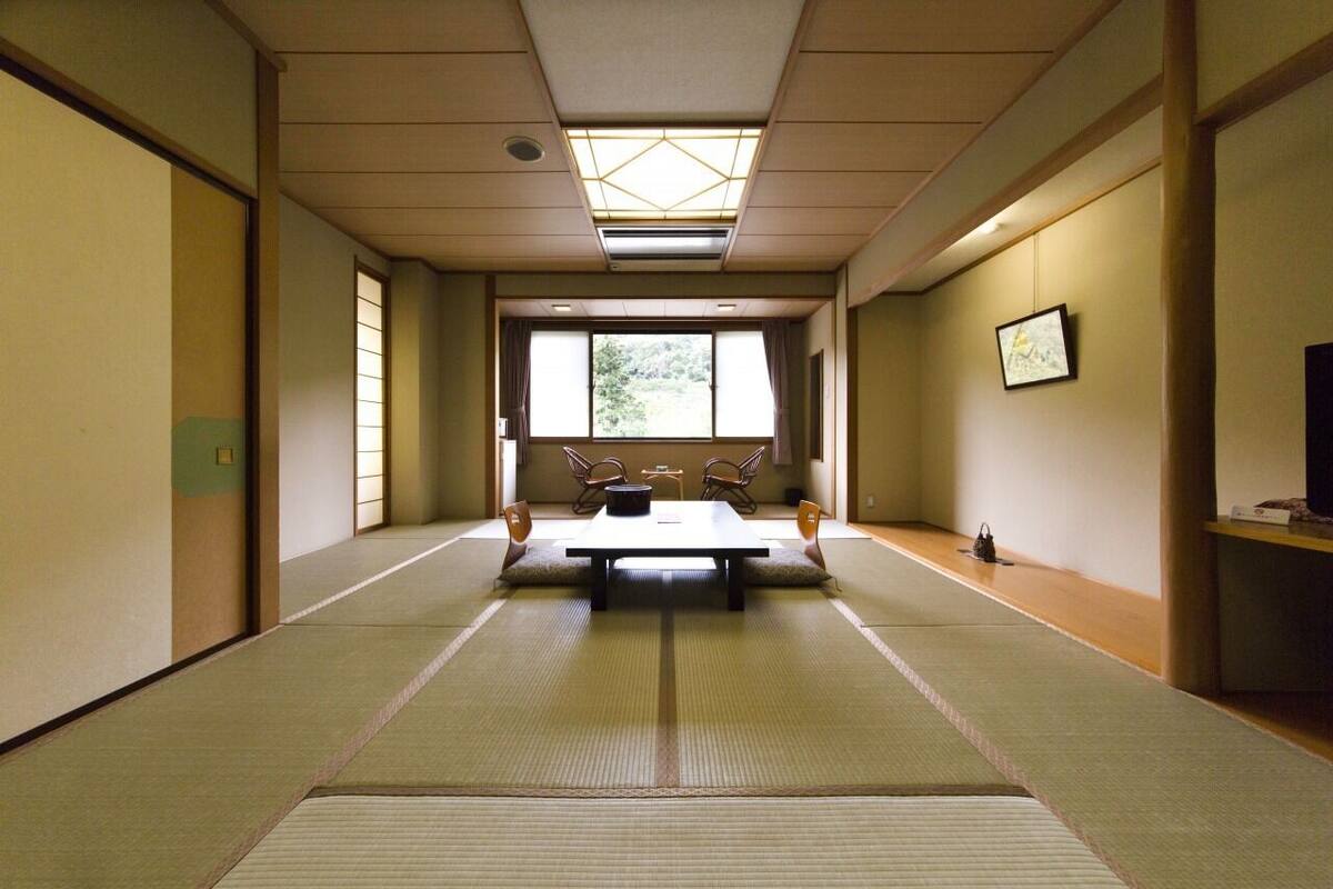 A traditional Japanese room with tatami flooring, a low table with chairs, a window with a view of greenery, and a ceiling fan.